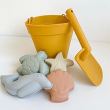 Load image into Gallery viewer, the silicone moulds in a green turtle, tan starfish, blue dolphin, and peach seashell in front of a orange bucket and spade