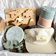 Load image into Gallery viewer, a gift box displaying a wooden dinosaur puzzle, silicone stacking cups, a crew cap in cream colour, and a collapsed snacking cup and easy grip spoon and fork in sage colour
