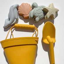 Load image into Gallery viewer, the silicone moulds in a blue dolphin, peach seashell, green turtle and tan startfish laying flat above the orange bucket and spade