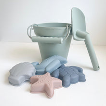 Load image into Gallery viewer, the silicone moulds in a grey seashell, pink starfish, blue dolphin, and blue turtle in front of a green bucket and spade