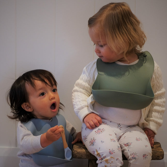 a child wearing a baby blue silicone bib holding a baby blue beechwood spoon with another child wearing a sage silicone bib