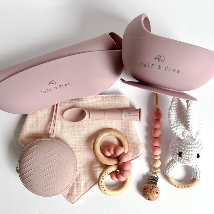 Folded silicone bib and suction bowl, soother case and spoon in rose colour, a wooden silicone ring teether, a silicone dummy clip in blush colour,  a white crochet bunny rattle with a baby pink muslin bib
