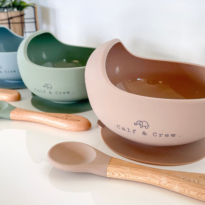 three silicone suction bowls in baby blue, sage and latte, with their matching beechwood spoons in front of them