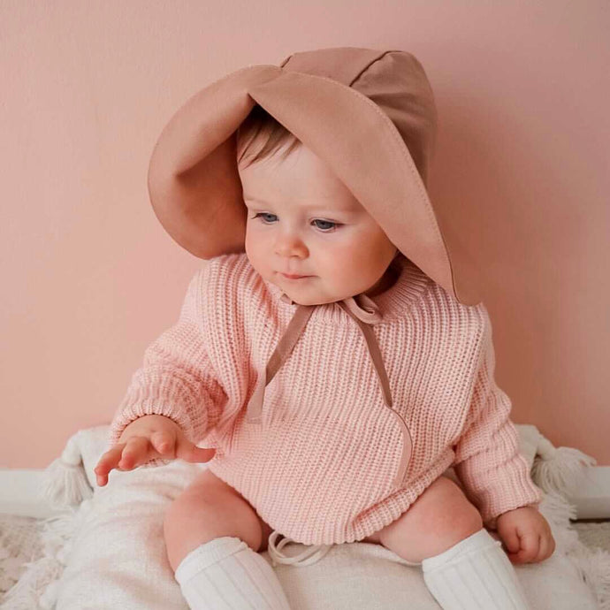 Baby sitting against a pink wall wearing the floppy sun hat in rose