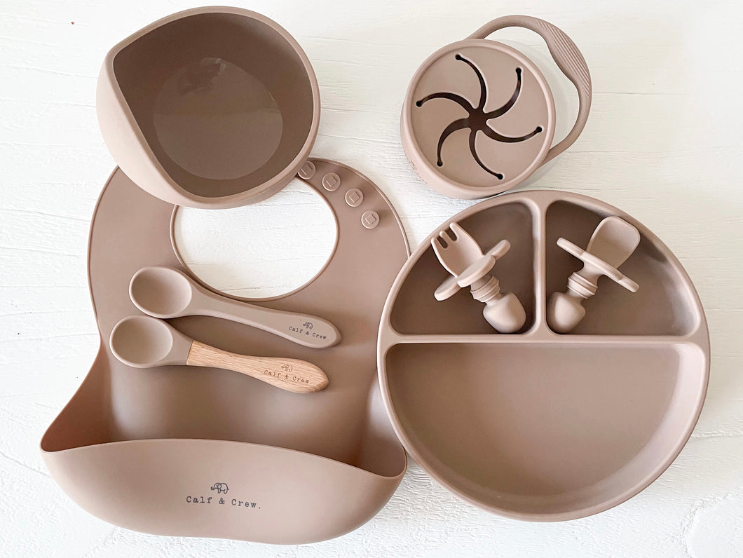 a suction bowl, a bib, a silicone spoon, a beechwood spoon, a collapsible snack cup, a suction divider plate, and easy grip cutlery all in latte silicone colour