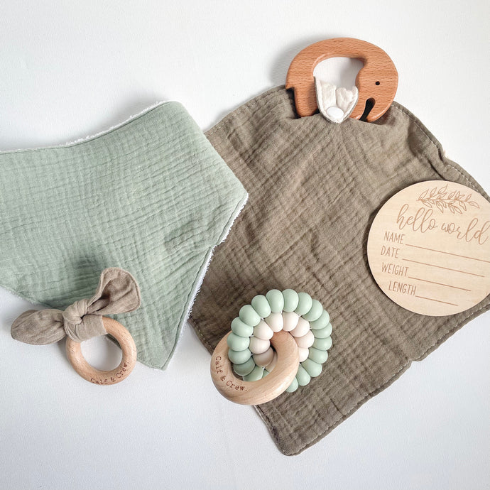Sage coloured muslin towel dribble bib with muslin bunny ear teether in taupe on a dark olive muslin comforter attached to a wooden elephant teether, a double silicone ring teether in mint and a 