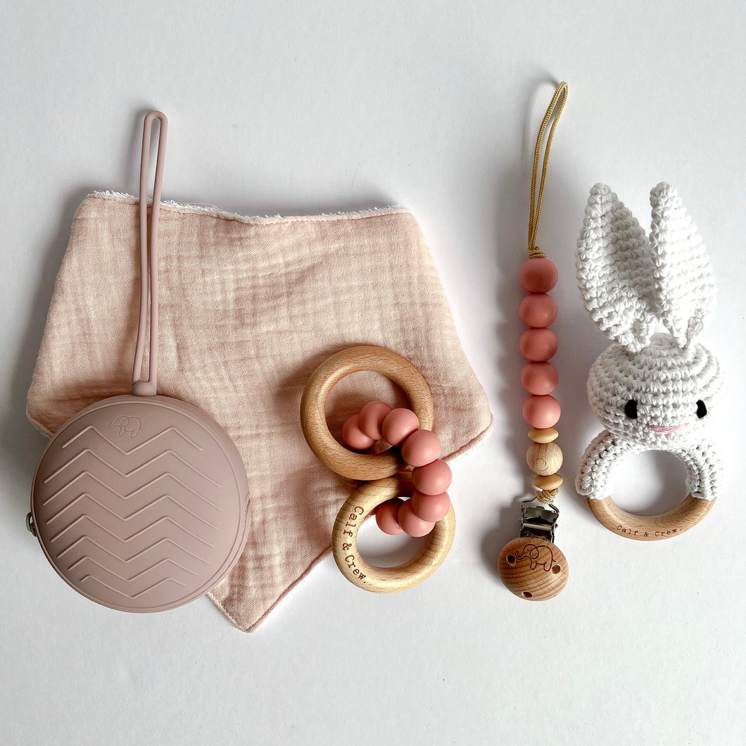 Baby pink coloured muslin bib with white crochet bunny rattle, wooden silicone ring teether, silicone dummy clip in blush colour and soother case in rose colour