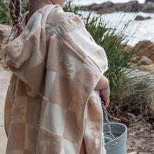 Load image into Gallery viewer, a child wearing the hooded terry beach towel carrying a bucket with the beach in the background
