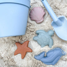 Load image into Gallery viewer, blue bucket and spade with the silicone shape moulds all laying in the sand
