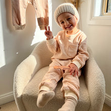 Load image into Gallery viewer, a child sitting in a soft white chair smiling wearing the tracksuit romper in peach striped
