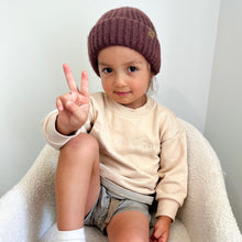 Load image into Gallery viewer, a child sitting in a soft white chair wearing the crew sweatshirt in eggnog and toddler beanie in chestnut
