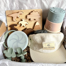 Load image into Gallery viewer, a gift box displaying a wooden dinosaur puzzle, silicone stacking cups, a crew cap in cream colour, and a collapsed snacking cup and easy grip spoon and fork in sage colour
