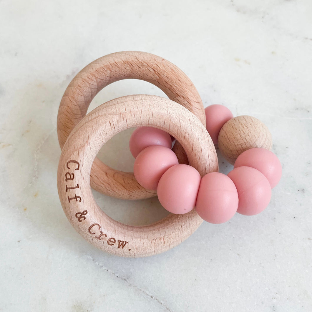 a wooden teether with a blush silicone ring and the calf & crew logo engraved on it