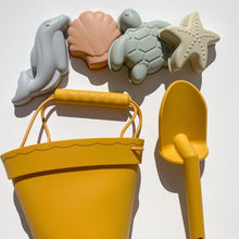 Load image into Gallery viewer, the silicone moulds in a blue dolphin, peach seashell, green turtle and tan startfish laying flat above the orange bucket and spade
