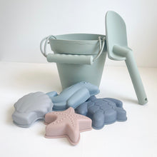 Load image into Gallery viewer, the silicone moulds in a grey seashell, pink starfish, blue dolphin, and blue turtle in front of a green bucket and spade
