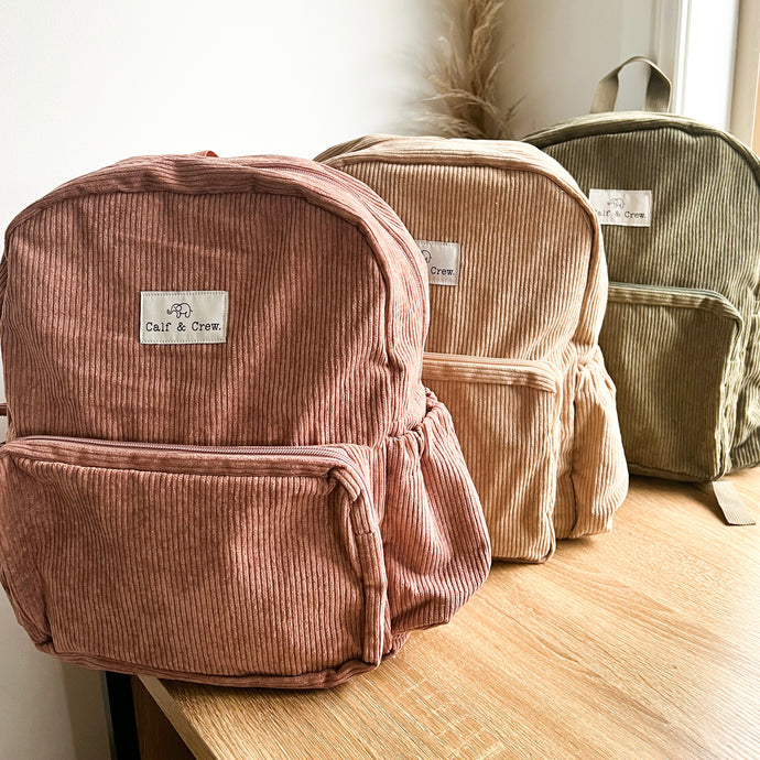 three corduroy junior backpacks in dusty rose, sand and khaki sitting on a wooden table