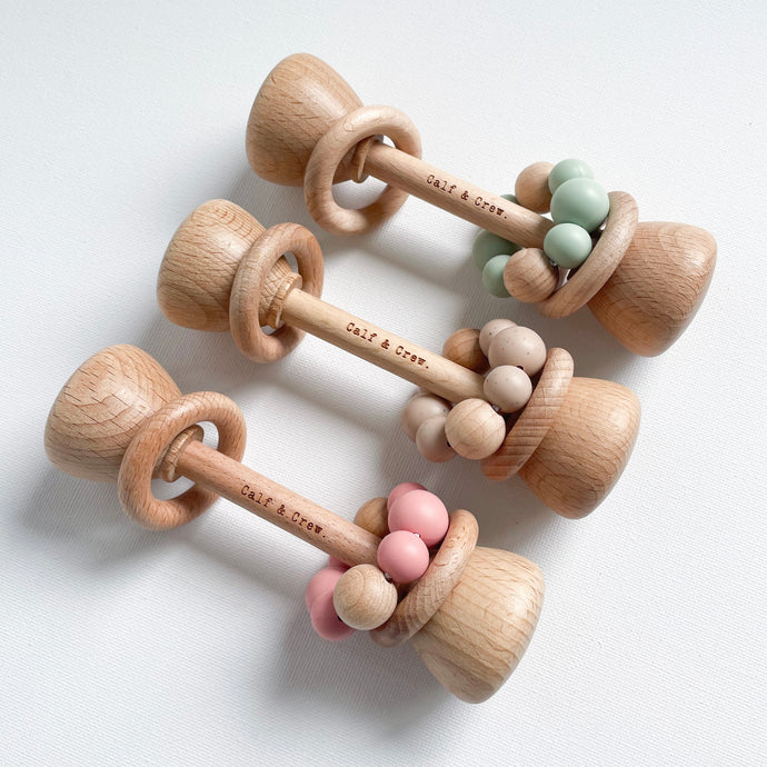 three wooden silicone rattles lined up in blush, chai, and mint colours