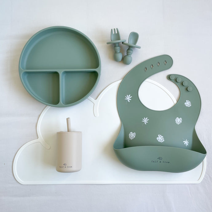 Divided plate, small fork and spoon, and silicone bib in forest green, laying on a white mat with a silicone straw cup in sand colour
