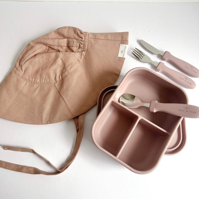 A folded floppy sun hat in rose colour with a open silicone bento lunchbox in rose and stainless steel spoon, fork and knife in rose colour